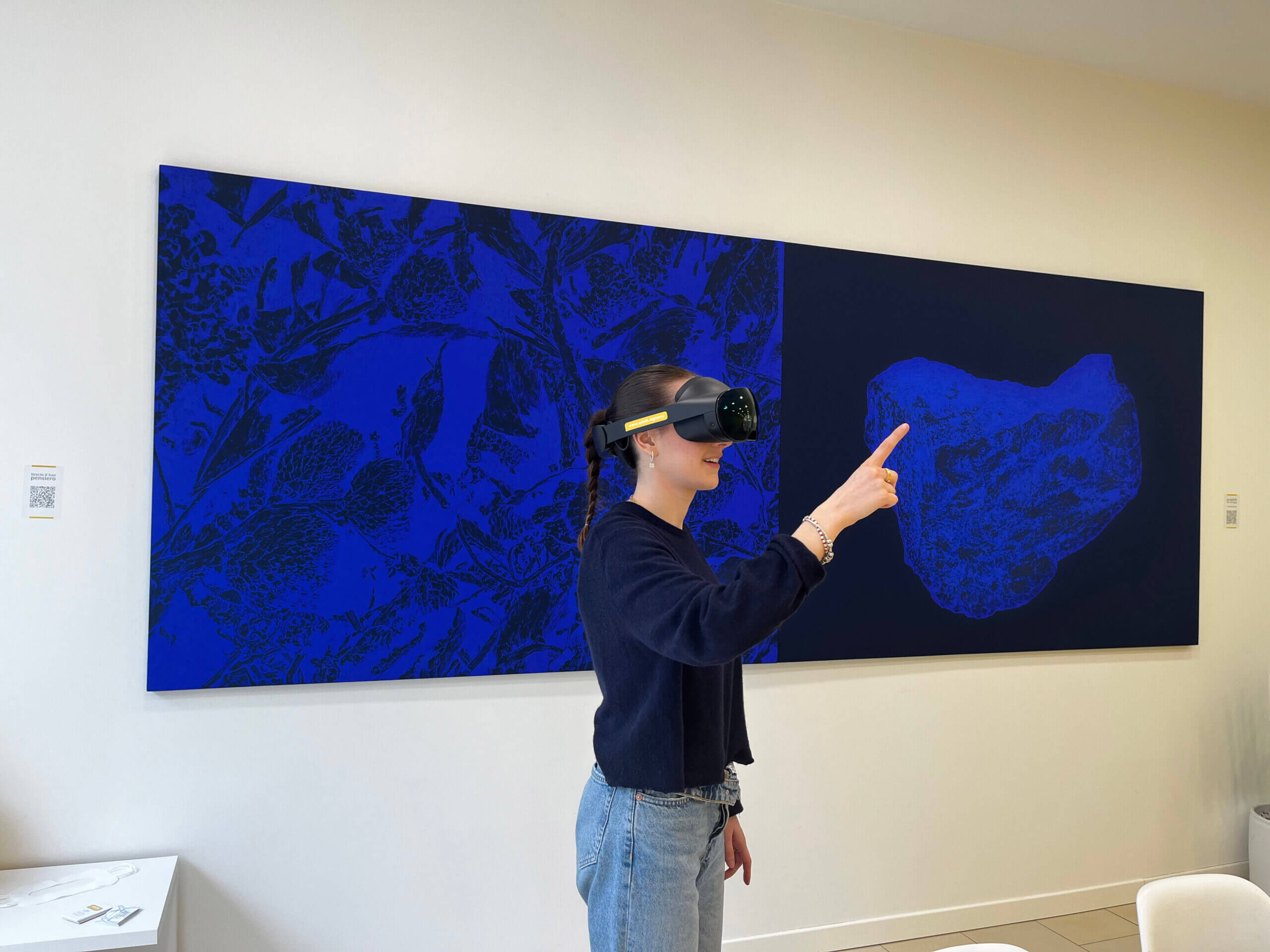 Una ragazza con indosso un visore per la realtà virtuale indica il cielo con alle spalle un quadro blu.
